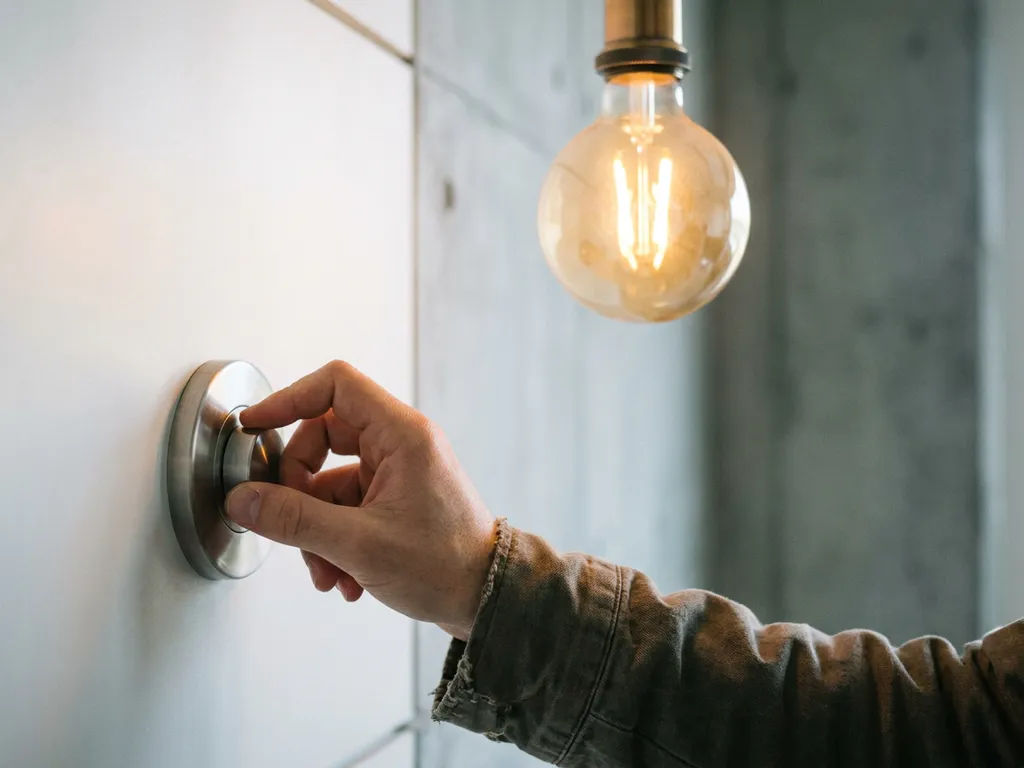 Hand die een witte LED-dimmer draait in modern interieur, met warm gloeiende LED-lamp op de achtergrond.